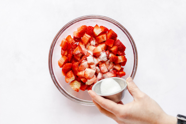 Image, taken from above, shows a hand sprinkling cornstarch over a large glass bowl of cut strawberries. 