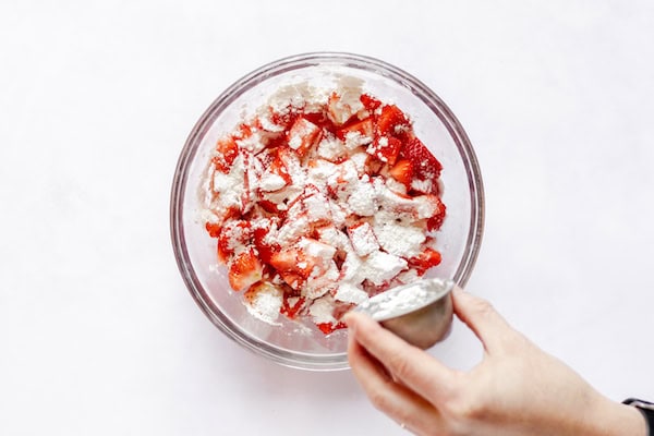 Image, taken from above, shows a hand sprinkling sugar over a large glass bowl of cut strawberries. 