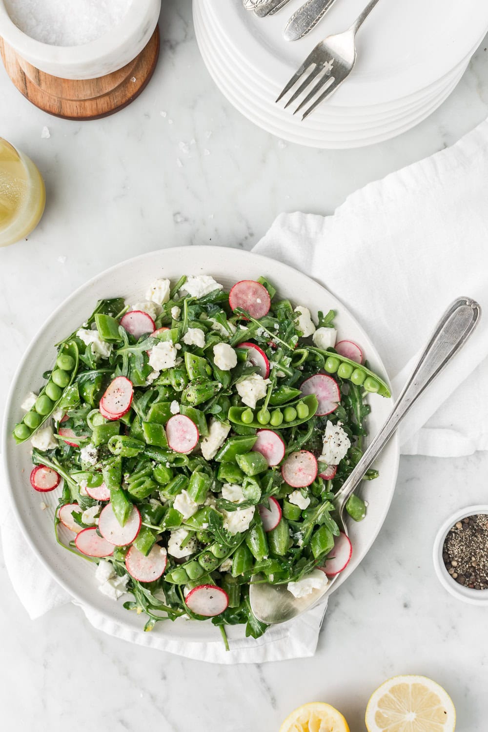 Image shows a white plate on a marble table full of a green, snap pea salad. The salad contains snap peas, radishes, feta and arugula.  