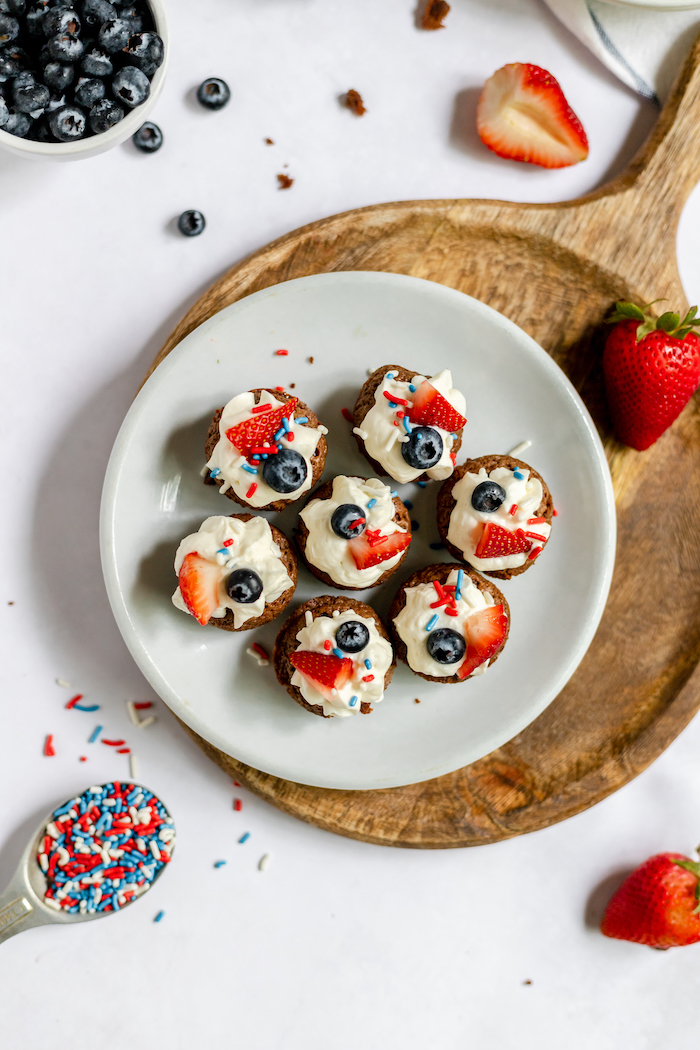Image, taken from above, shows a white plate with small brownie bites with frosting, blueberry and strawberry on top. The plate rests on a wooden tray, with blueberries and strawberries nearby.
