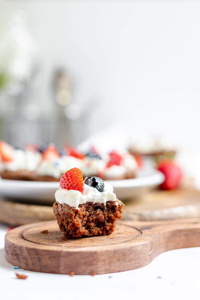 Image shows a single gluten free brownie bite with frosting, blueberry and strawberry on a wood tray. A plate of more brownie bites sit on a plate in the background.