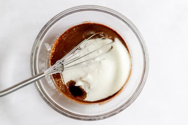 Image, taken from above, shows a glass bowl with melted chocolate and butter, with a whisk adding the sugar in.