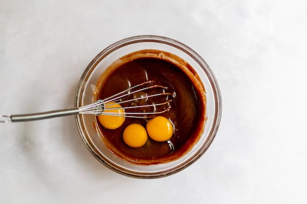 Image, taken from above, shows a glass bowl with melted chocolate and a whisk adding eggs to the batter.