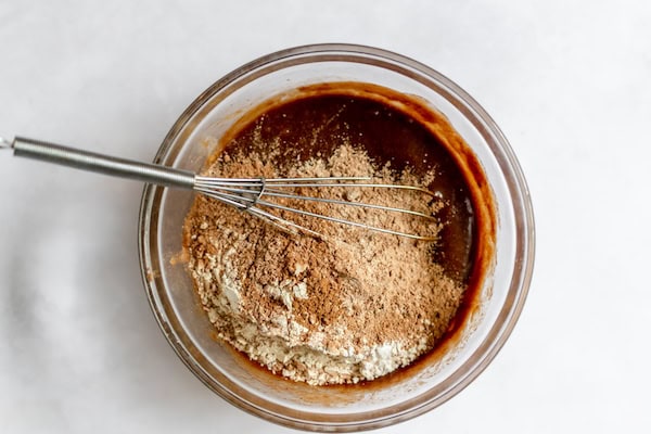 Image, taken from above, shows a glass bowl with batter for gluten free brownie bites, with a whisk and dry ingredients ready to be whisked in.