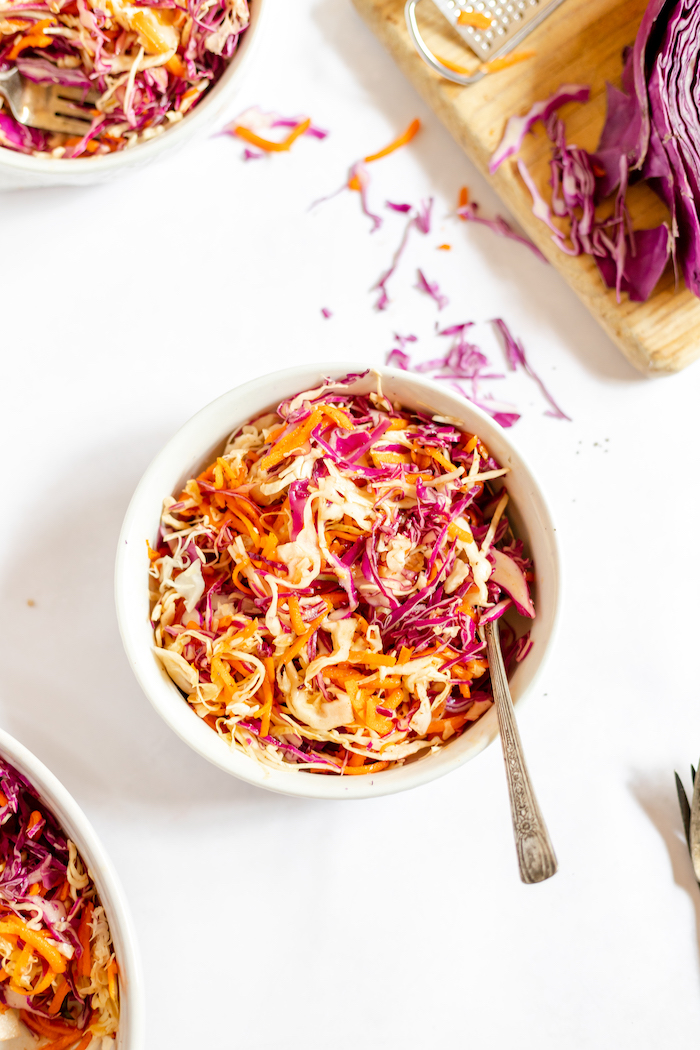 Image shows a white bowl full of coleslaw made with purple cabbage and carrots, without mayo. A cutting board with purple cabbage sits near the bowl.