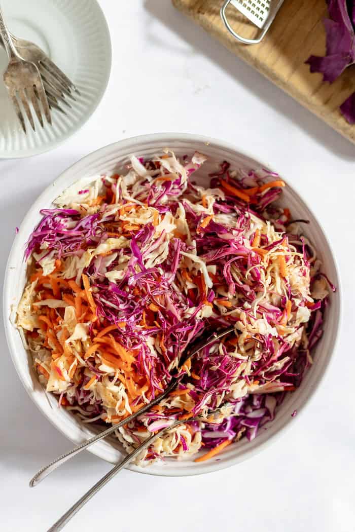 Image shows a large bowl of coleslaw without mayo, photographed from above, sitting on a table. Nearby is a white plate with some forks on it.