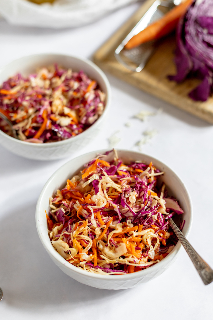 Image shows two white bowls full of coleslaw made from purple cabbage and carrots sitting on a white table. A cutting board with a carrot and cabbage sits in the background.
