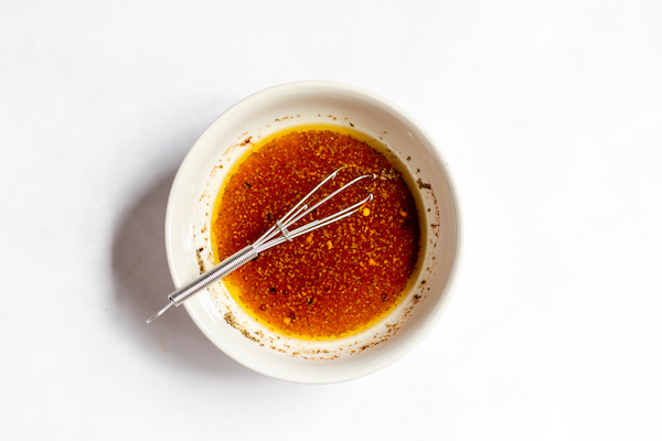 Image shows a white bowl photographed from above with a whisk in it. The bowl is full of oil and seasoning mixed in for coleslaw without mayo