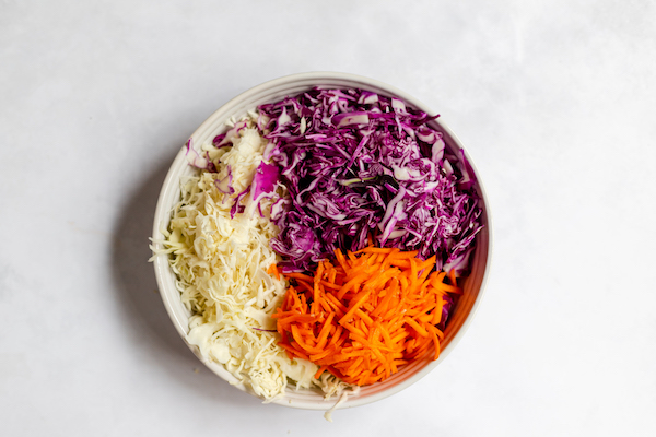 Image shows a photo taken from above of a white bowl full of ingredients to make coleslaw. The purple and white cabbage, and orange carrots each take up about one third of the bowl. 