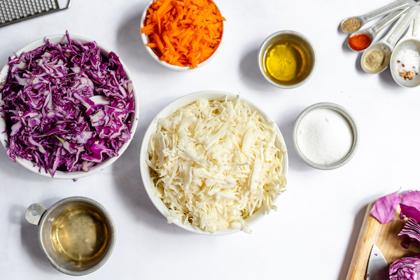 Photo shows a white table with several bowls on it. Each bowl is filled with different ingredients to make coleslaw without mayo- cabbage, carrots, oils and seasonings.