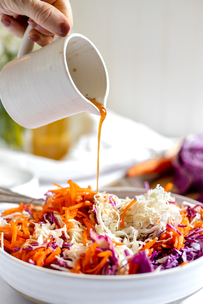 Photo shows a hand holding a small white pitcher, pouring an oil and seasoning mixture over a large bowl of cabbage and carrots to make coleslaw without mayo