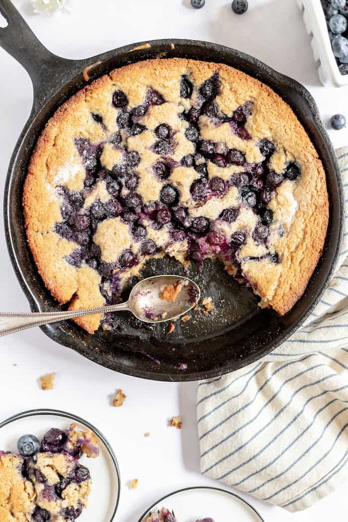 Image, taken from above, shows a gluten free blueberry cobbler in a cast iron skillet. About 1/4 of the cobbler has been scooped away with a spoon, still resting in the skillet. 