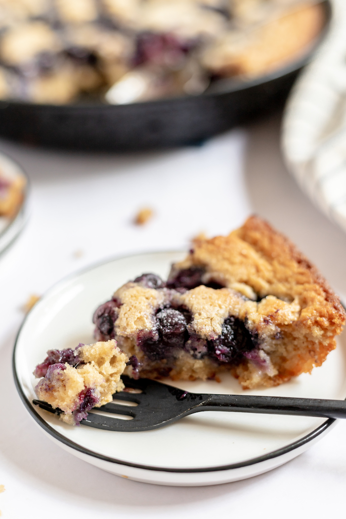Photo shows a white plate with a slice of blueberry cobbler on it. A fork resting on the plate holds a small bite of cobbler.