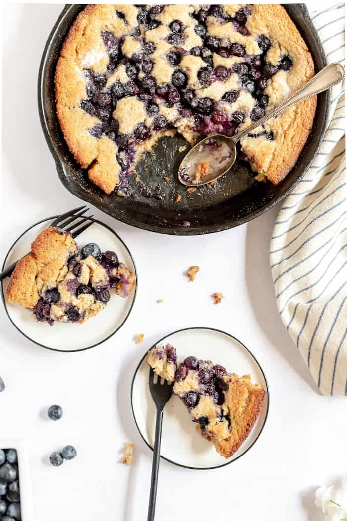 Photo, taken from above, shows two small white plates with black trim, each with a piece of blueberry cobbler on it. Next to the plates is a cast iron skillet with the rest of the cobbler in it.  