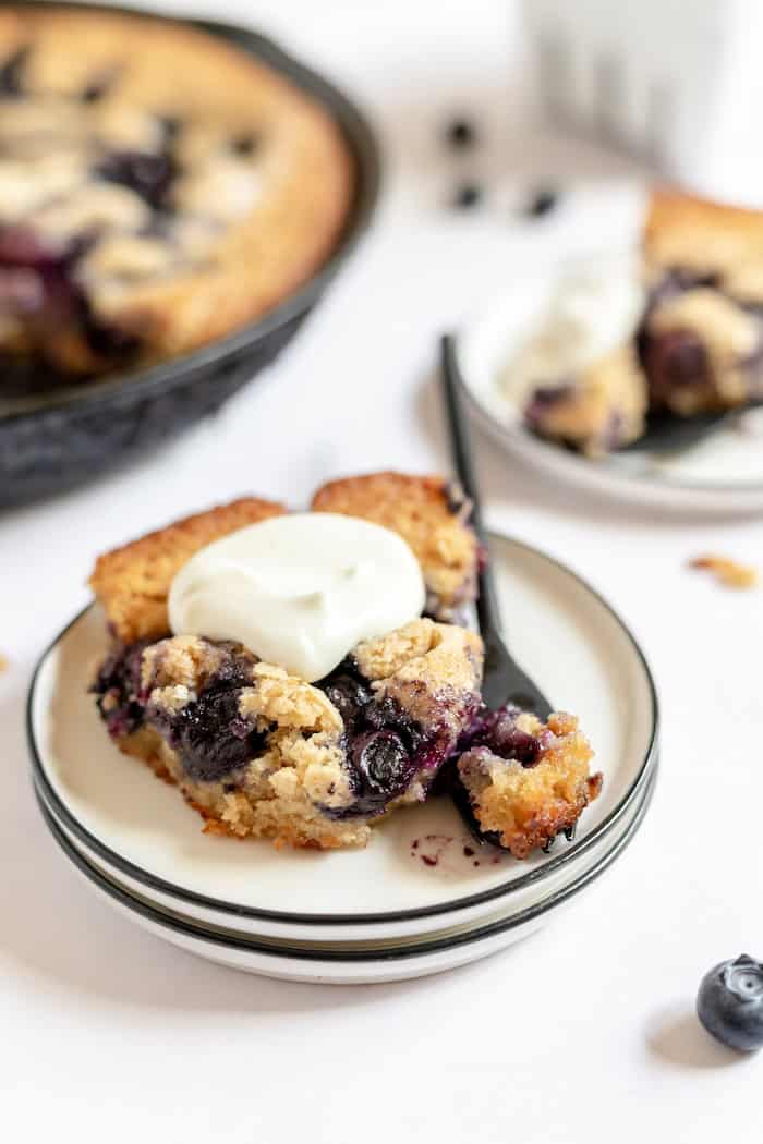 Image shows a plate with a piece of blueberry cobbler with homemade whipped cream on top and a fork resting with it. In the background is a skillet of more cobbler. 