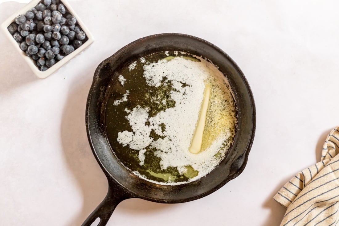 Image shows a cast iron skillet with melter butter and a bowl of blueberries sitting nearby.
