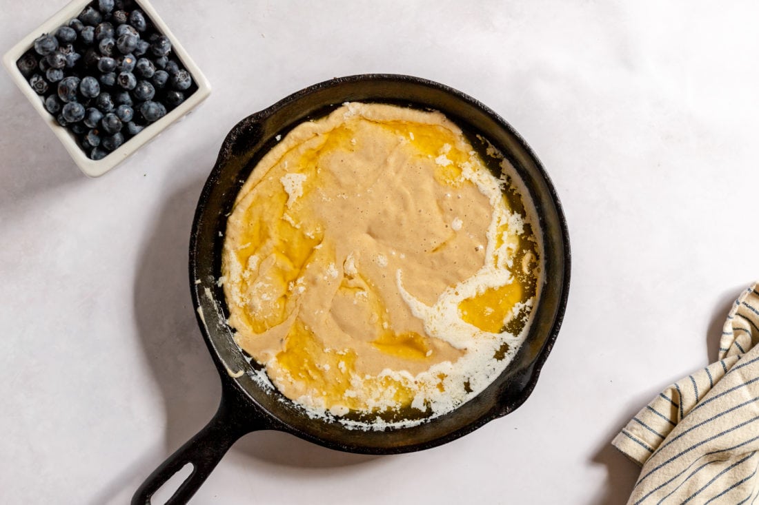 Image, taken from above shows a cast iron skillet with melted butter and other ingredients for a blueberry cobbler. A square bowl of bluberries sits next to the skillet. 