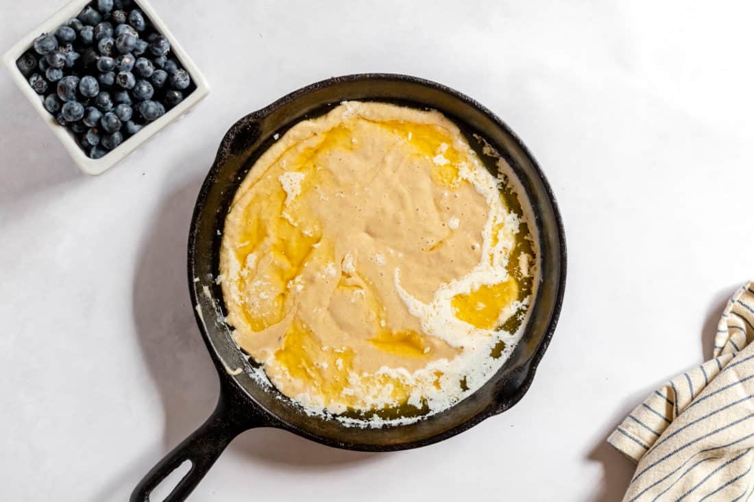 Photo, taken from above, shows a cast iron skillet with flour, sugar, and butter mixed in melted butter to make blueberry cobbler. A square bowl of blueberries sits next to the skillet. 