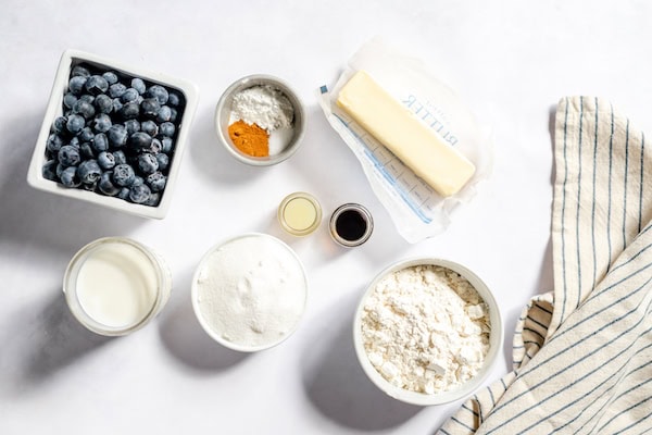 Image, taken from above, shows bowls on a white table holding all the ingredients for blueberry cobbler. Bowls contain blueberries, flour, sugar, butter, and other seasonings. 