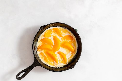 Image shows an iron skillet with the peach cobbler ingredients with peaches in it.
