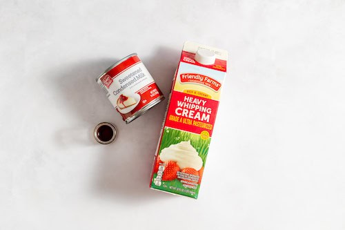 Image shows a carton of heavy whipping cream, a jar of sweetened condensed milk, and a small teaspoon of vanilla on a white table.