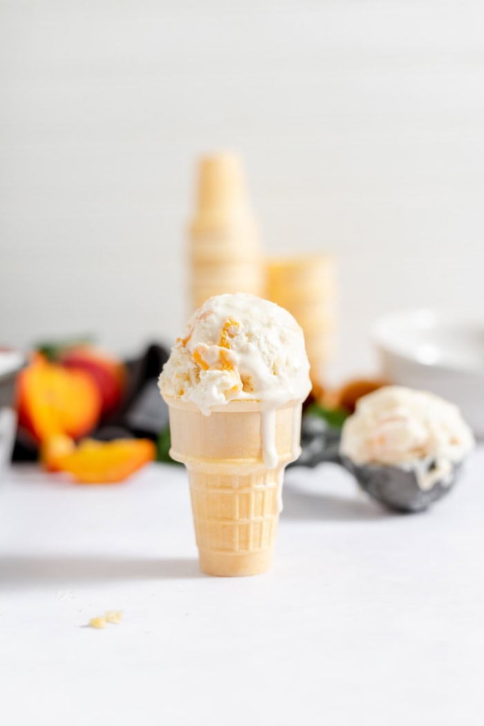 Image shows an ice cream cone set on a white table, with a scoop of peach cobbler ice cream melting down the sides. In the background are sliced peaches, and a scoop of ice cream. 