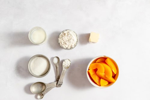 Image shows a white table with bowls of peaches, flour, sugar, and butter set out to make the peach cobbler ice cream.