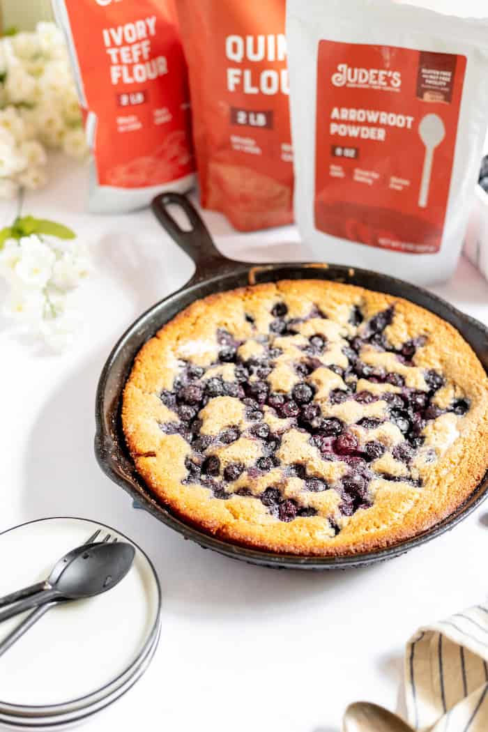 Image shows a cast iron skillet with a blueberry cobbler in it. Behind it are three bags of Judee's ivory teff flour, quinoa flour, and arrowroot powder to make the cobbler.