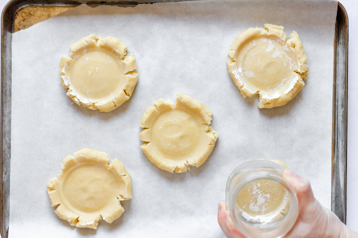 Image, taken from above, shows five sugar cookies with a hand pressing a glass to flatten them. 