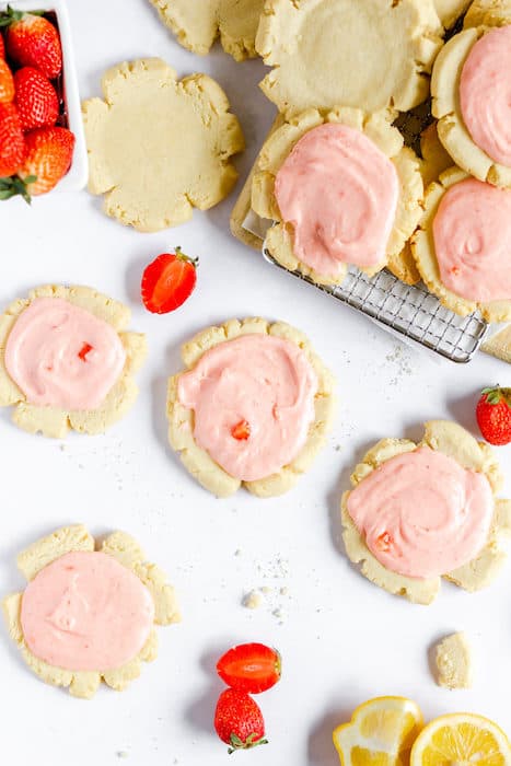 Image shows gluten free sugar cookies placed on a cooling rack, and also a counter top. Some of the cookies are iced with pink, strawberry icing. A few sliced strawberries are lying near the cookies on the counter. 