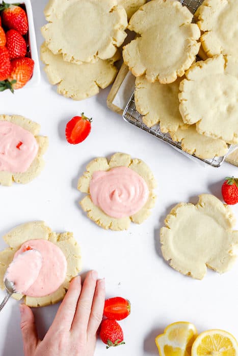 Image shows gluten free sugar cookies placed on a cooling rack, and also a counter top. Some of the cookies are iced with pink, strawberry icing. A few sliced strawberries are lying near the cookies on the counter. 