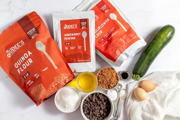 Image shows the ingredients displayed to make gluten free chocolate zucchini bread- quinoi flour, arrowroot powder, cauliflower powder, a zucchini, eggs, chocolate chips, oil, sugar, and more