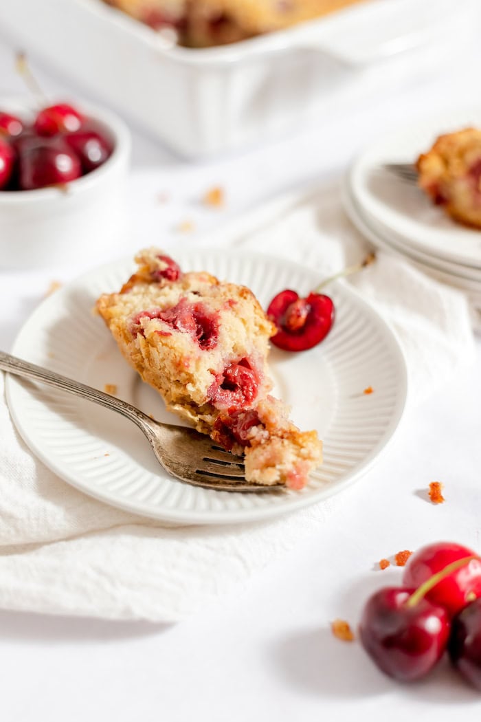 Image shows a small plate of sour cherry cake with a fork resting on it. In the background is a casserole dish with the rest of the cake, and a small bowl of cherries. 