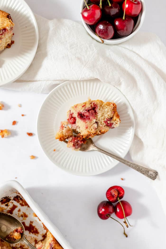 Photo, taken from above, shows a white plate with a piece of sour cherry cake sitting on it with a fork. Nearby is a small bowl of cherries, and another plate of cake. 