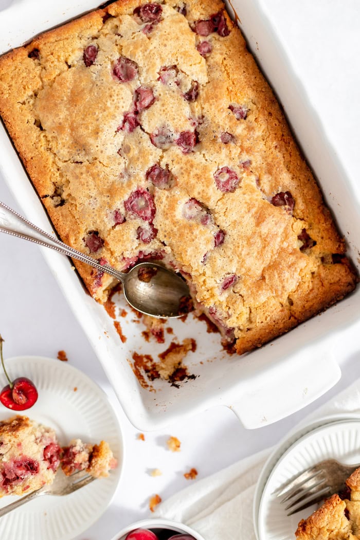 Image, taken from above, shows sour cherry cake in a white pan, with a spoon resting where one piece has been removed. 