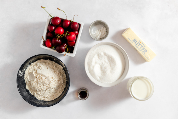 Photo shows all the ingredients needed to make sour cherry cake- a bowl of flour and sugar, a container of cherries, vanilla and butter