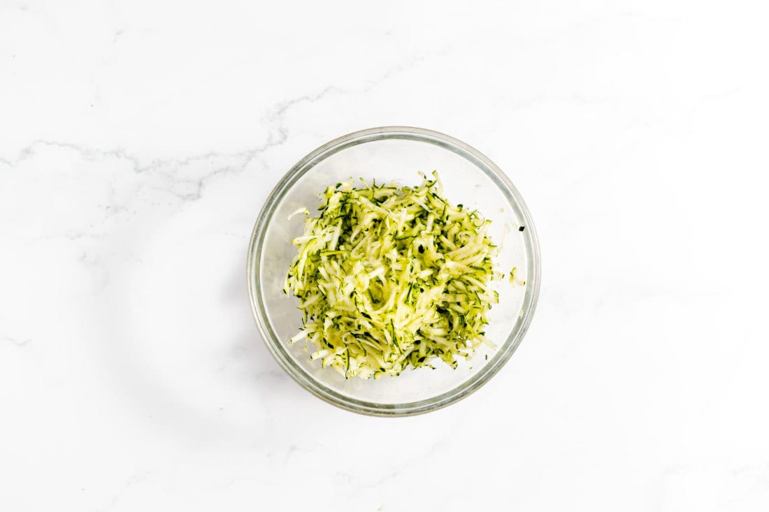Image shows a glass bowl on a white countertop with shredded zucchini in it.