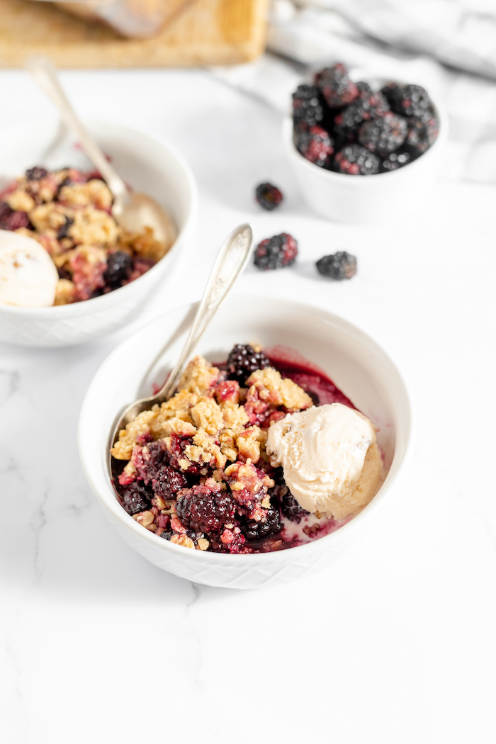 Image shows a counter top with two bowls of gluten free blackberry crisp with a scoop of vanilla ice cream. Nearby sits a small bowl of blackberries, with three laying next to the bowl on the countertop. 