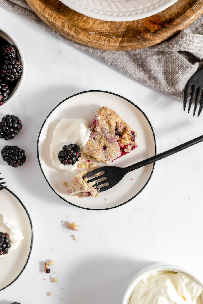 Image, taken from above, shows a small white plate with black trim with a blackberry scone on it. A fork with a bite of scone rests next to the scone.