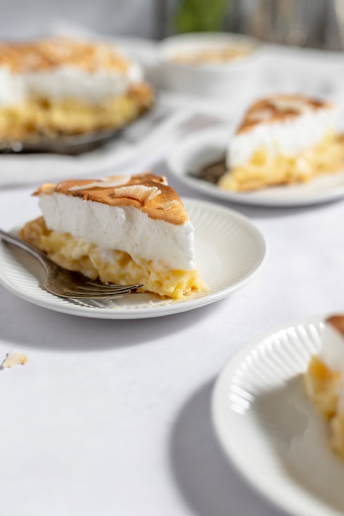 Image shows several plates on a white table holding slices of coconut meringue pie. The plate in the foreground has a fork resting on the side of it. 