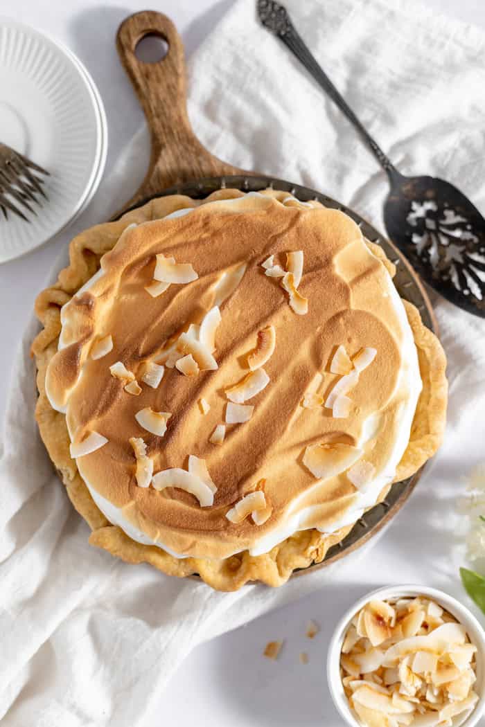Image, taken from above, shows a complete coconut meringue pie, topped with coconut chips, sitting on a white tablecloth. 