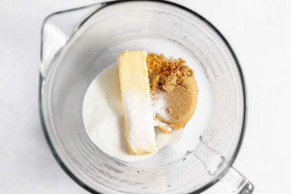 Image shows a large glass mixing bowl with sugars and butter ready to be blended for peanut butter cookies