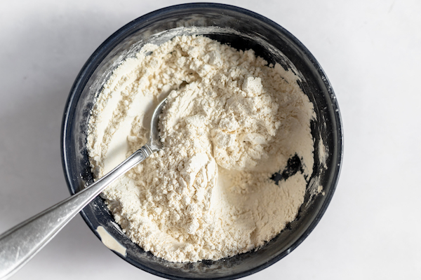 Image shows a black mixing bowl with flour and other dry ingredients mixed with a spoon