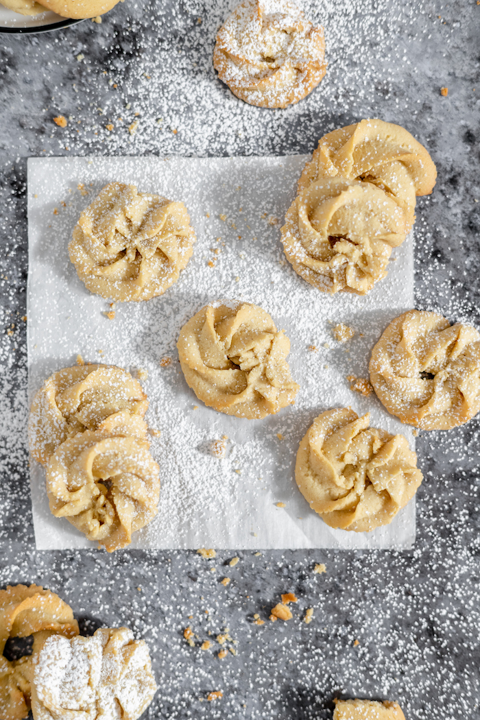 Image, taken from above, shows gluten free butter cookies on a sheet of parchment, sprinkled in powdered sugar.