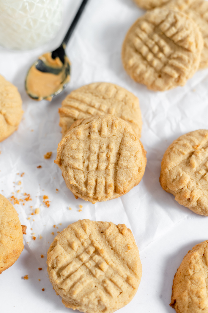 Image shows a half dozen or so gluten free peanut butter cookies spread out on parchment paper. Nearby sits a spoon with peanut butter on it. 