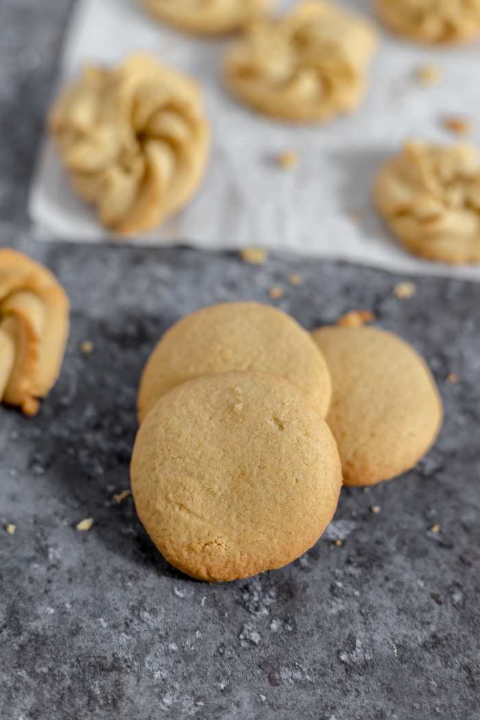 Image shows several round gluten free butter cookies stacked on a counter. In the background is a piece of parchment paper with more cookies cooling.