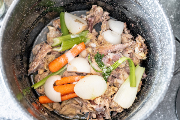 Image, taken from above, shows a large kettle full of chicken bones and vegetables to make broth.