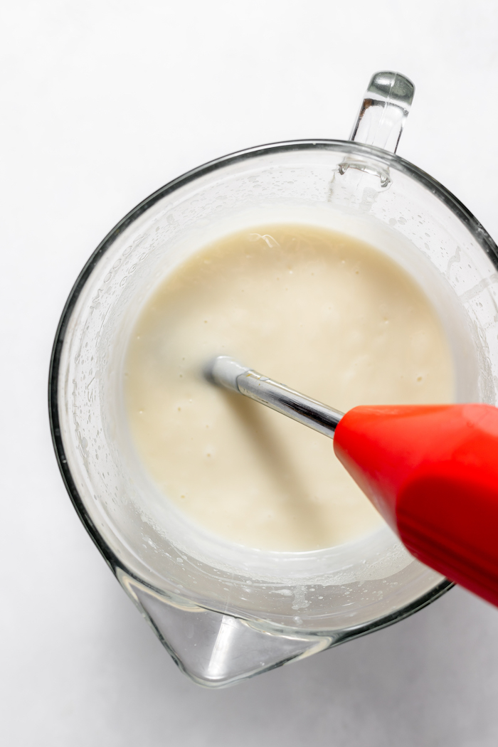 Photo shows an immersion blender in the soap mixture in a glass measuring cup, photographed from above,  blending at a low pace