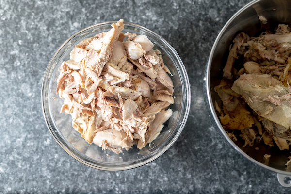 Photo, taken from above, shows a glass bowl full of chicken pieces, next to a stock pot of cooked chicken.