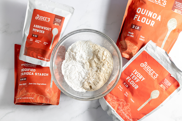 Image, taken from above, shows bags of Judee's products- Arrowroot powder, Tapioca starch, ivory teff flour and quinoa flour on a counter around a glass bowl of the flour mixture.